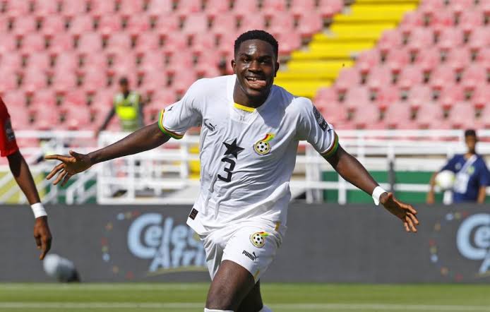 Soccer player in a white jersey (#3) celebrating with arms outstretched on a green pitch with stadium seats behind him, during a match.