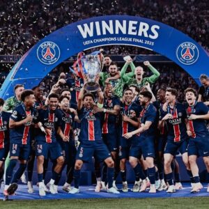 Paris Saint-Germain players celebrate winning the UEFA Champions League Final 2025, lifting the trophy under a blue 'WINNERS' arch with confetti.