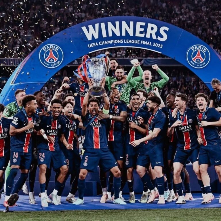 Paris Saint-Germain players celebrate winning the UEFA Champions League Final 2025, lifting the trophy under a blue 'WINNERS' arch with confetti.