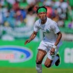 Soccer player in white kit with a green headband sprinting on a field with a blurred crowd in the background.