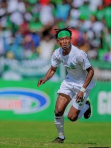 Soccer player in white kit with a green headband sprinting on a field with a blurred crowd in the background.