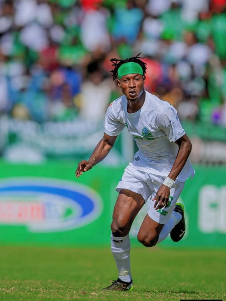 Soccer player in white kit with a green headband sprinting on a field with a blurred crowd in the background.