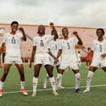 Four female soccer players in white uniforms celebrate with raised fists on a stadium field.