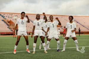 Four female soccer players in white uniforms celebrate with raised fists on a stadium field.