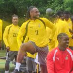 Group of soccer players in yellow training tops outdoors; a man with number 11 lifts his knee while others watch.
