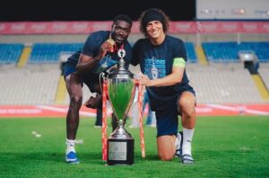 Two male athletes kneel on a football pitch, posing proudly with a large trophy between them.