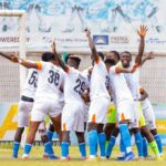 Soccer players in white jerseys celebrating in a huddle near the goal on a grassy field.