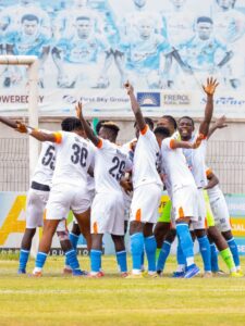 Soccer players in white jerseys celebrating in a huddle near the goal on a grassy field.