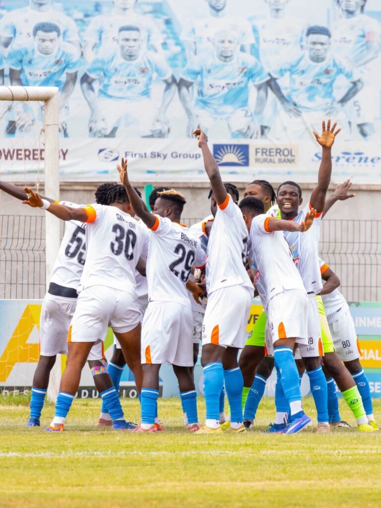 Soccer players in white jerseys celebrating in a huddle near the goal on a grassy field.