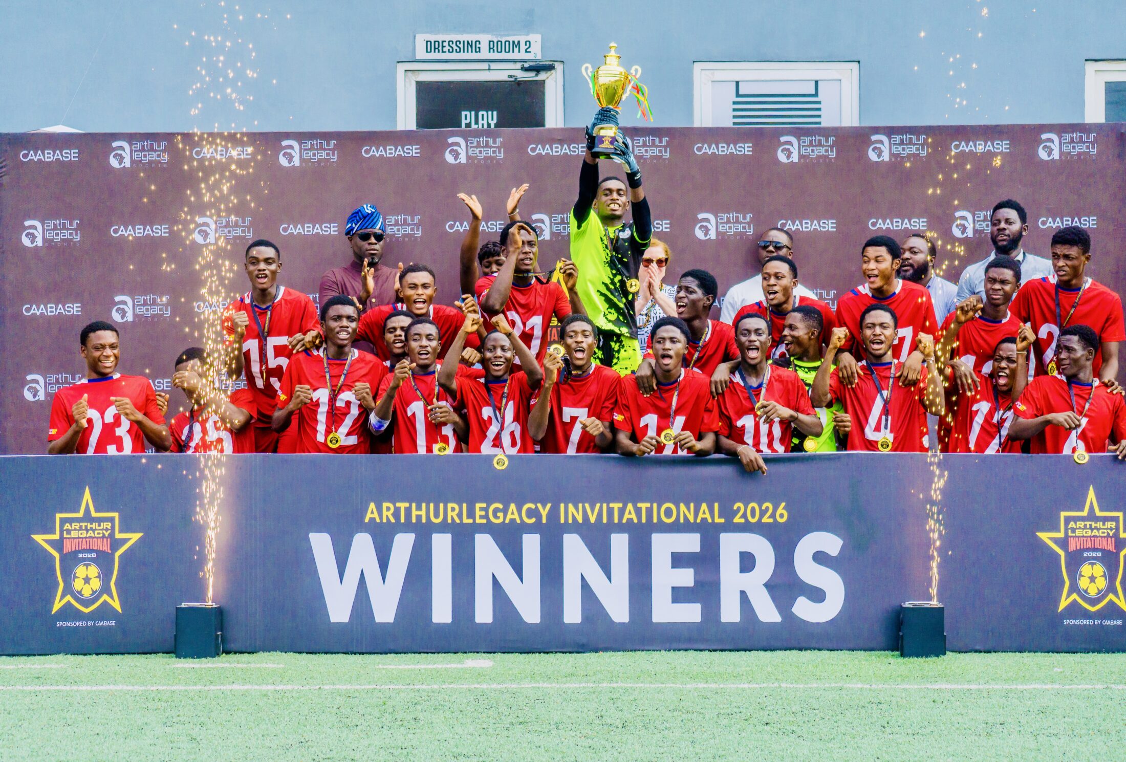 Group of young soccer players in red jerseys celebrate on stage, holding medals and a trophy aloft at an awards ceremony.