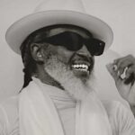 Man with a white beard and dark sunglasses, wearing a white hat and scarf, smiling against a light background.