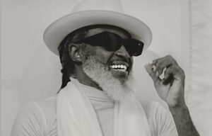 Man with a white beard and dark sunglasses, wearing a white hat and scarf, smiling against a light background.