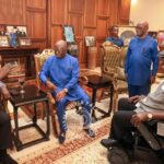 Group of men in a formal office, several in blue outfits, sit around a small table and talk; a man in a wheelchair joins the discussion while security personnel stand nearby.