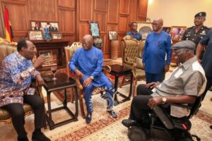 Group of men in a formal office, several in blue outfits, sit around a small table and talk; a man in a wheelchair joins the discussion while security personnel stand nearby.