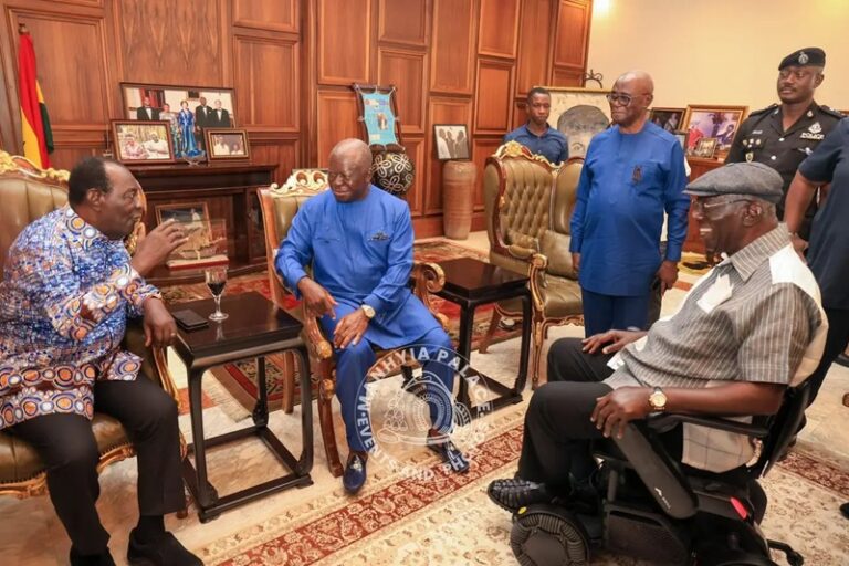 Group of men in a formal office, several in blue outfits, sit around a small table and talk; a man in a wheelchair joins the discussion while security personnel stand nearby.