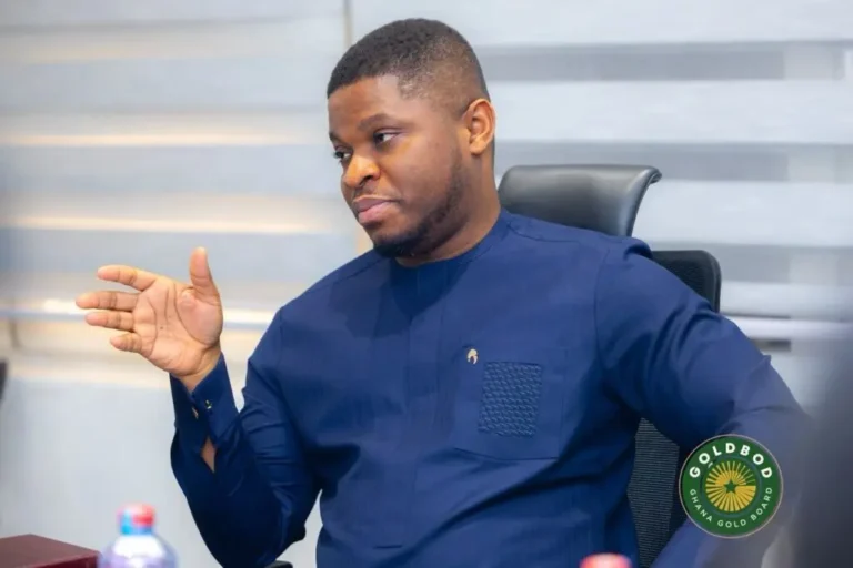 Man in a navy blue traditional shirt sits at a meeting table, gesturing with his left hand as he talks.
