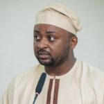 Man in a beige traditional outfit and cap speaking into a microphone