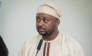 Man in a beige traditional outfit and cap speaking into a microphone