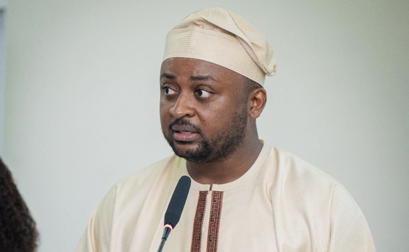 Man in a beige traditional outfit and cap speaking into a microphone