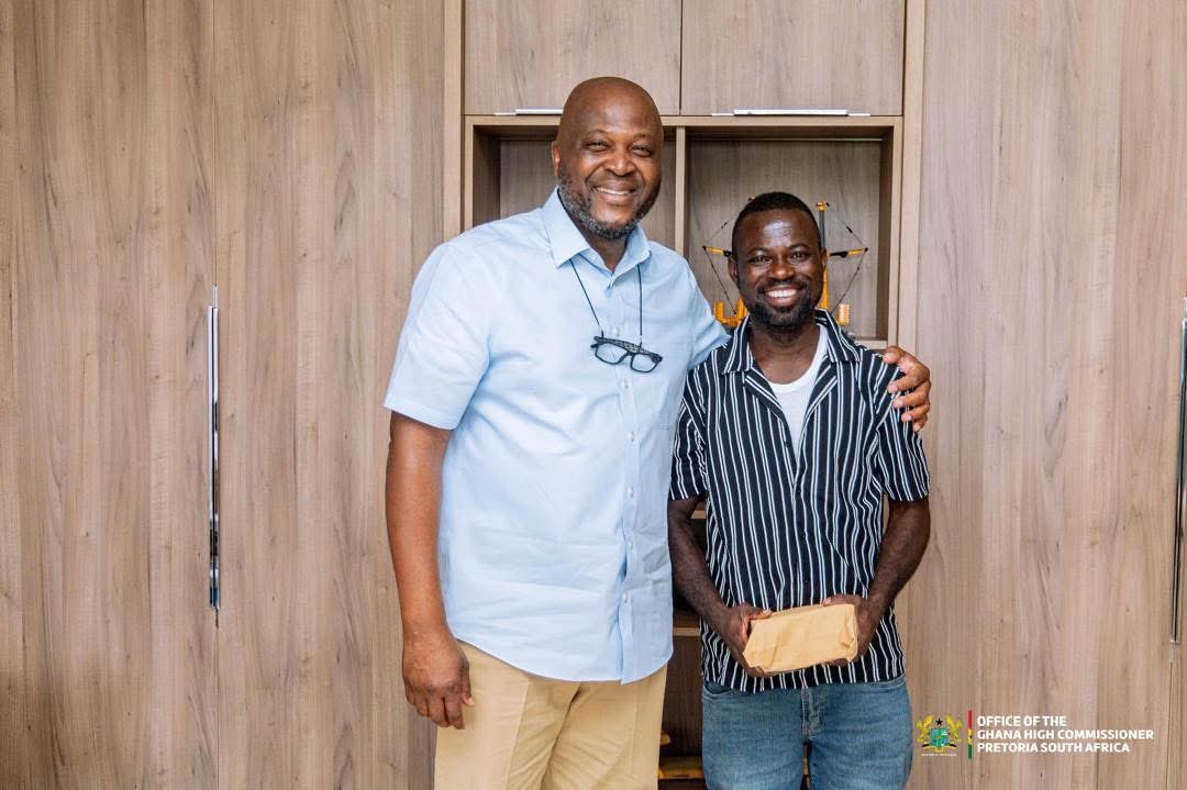 Two men smile and pose together in a wood-paneled room; the taller man wears a light blue shirt with glasses hanging from his neck, and has his arm around the other man.