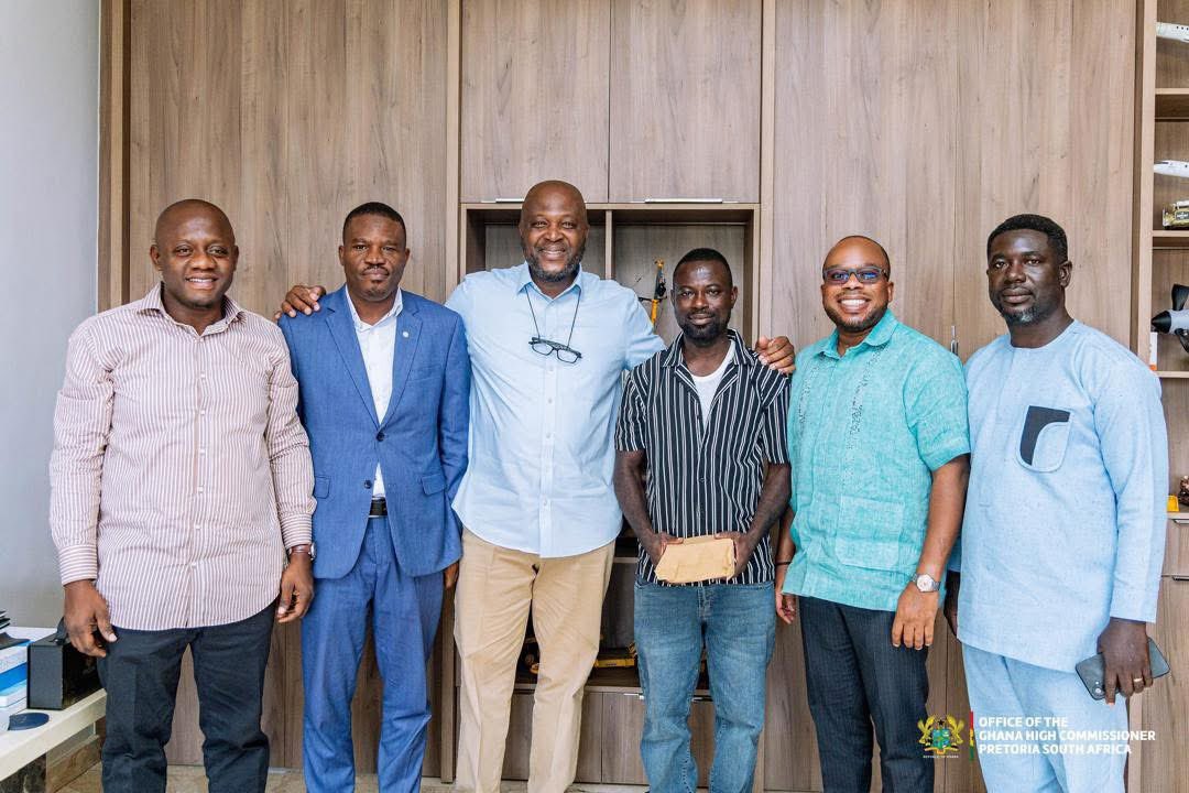 Six men stand side by side in an office, smiling, with wood-paneled cabinets behind them.