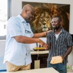 Two men in a bright office shake hands as one passes an envelope to the other, with a large lion portrait on the wall behind them.