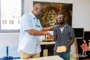 Two men in a bright office shake hands as one passes an envelope to the other, with a large lion portrait on the wall behind them.