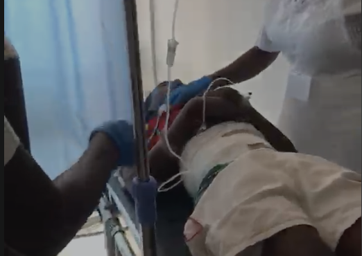 Two healthcare workers attend to a patient in a hospital bed, with IV lines and monitoring tubes visible.