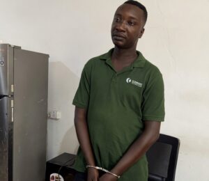 Man in a green polo stands with his hands cuffed in front, next to a chair in a small office area with a refrigerator nearby.