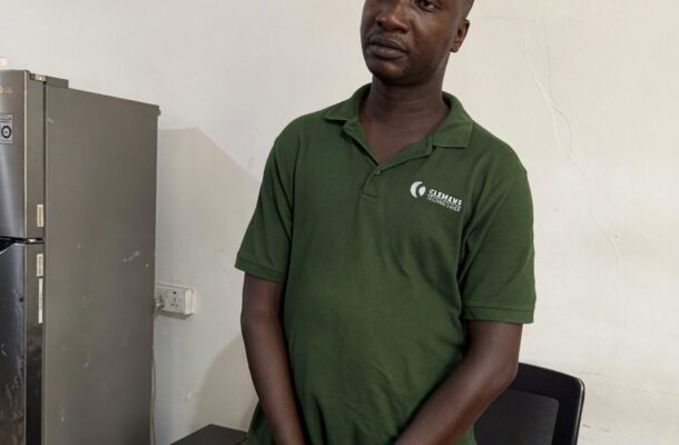 Man in a green polo stands with his hands cuffed in front, next to a chair in a small office area with a refrigerator nearby.