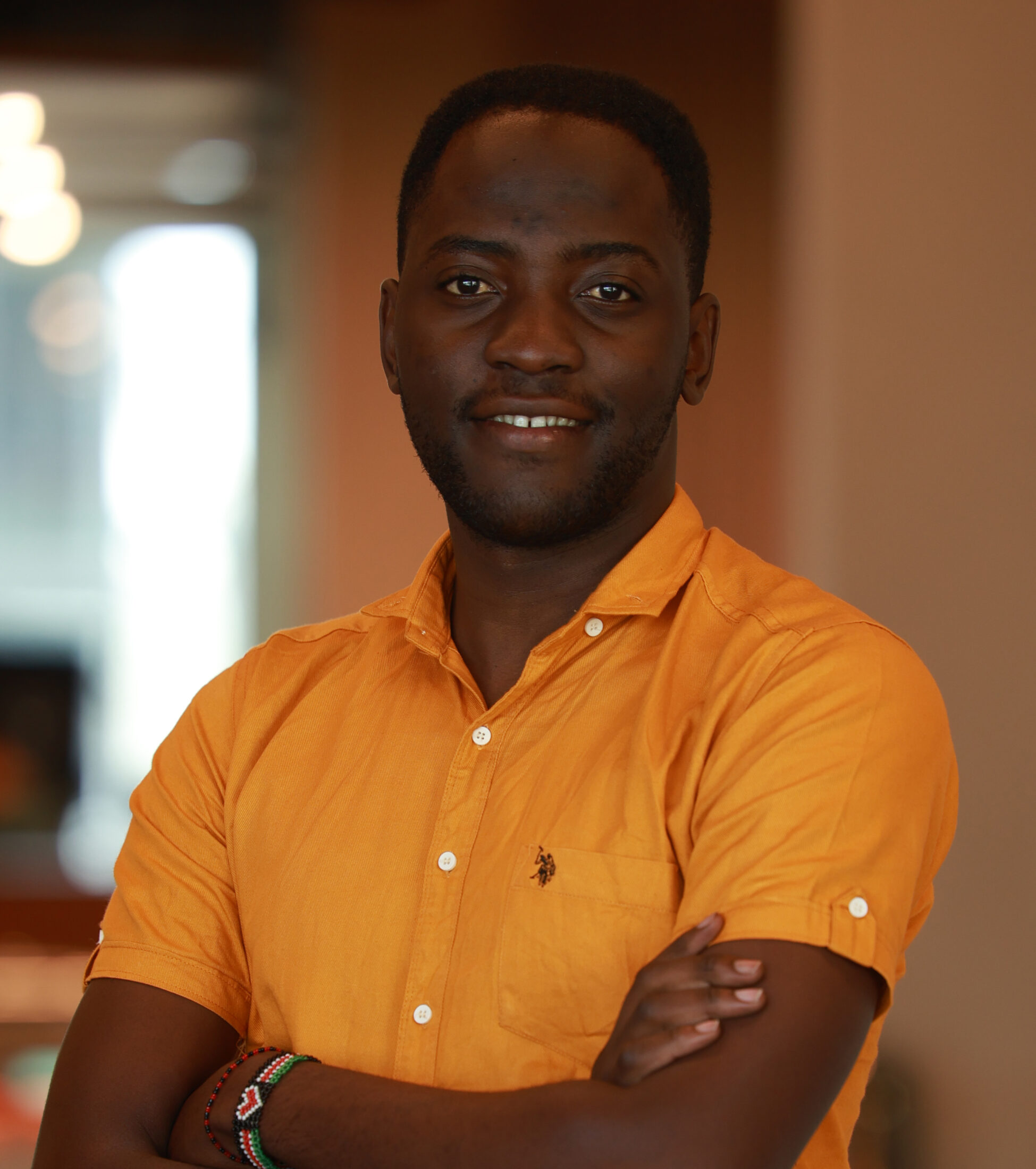 Portrait of a smiling man with dark skin wearing an orange shirt, arms crossed in a warm indoor setting.
