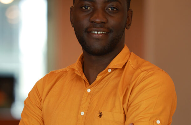 Portrait of a smiling man with dark skin wearing an orange shirt, arms crossed in a warm indoor setting.