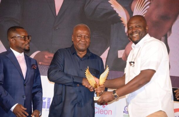 Three men on a stage at an awards event; center man accepts a golden winged trophy from a smiling recipient in a white shirt on the right, with a fourth man watching on the left.