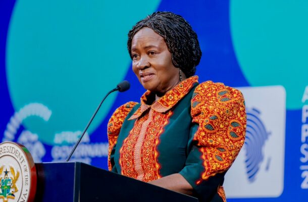 Woman wearing a red-and-gold patterned dress speaks at a podium with a microphone during a summit, seal on the podium visible against a blue-green backdrop.