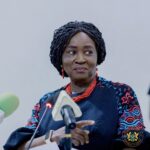Woman with braided hair in a patterned blue outfit and red jewelry stands at a podium, holding papers, with the Ghana presidential seal visible on her chest.
