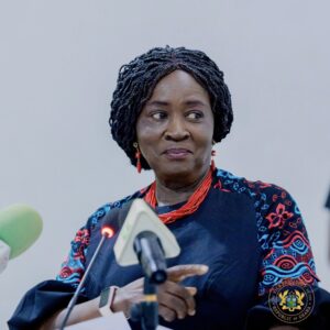 Woman with braided hair in a patterned blue outfit and red jewelry stands at a podium, holding papers, with the Ghana presidential seal visible on her chest.