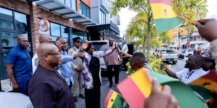 Group of people on a sidewalk greeting each other, several waving Ghana flags during a street gathering.