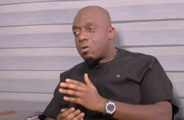 Man in a black shirt sits against a striped wall, gesturing with his hands as he speaks in an interview setting