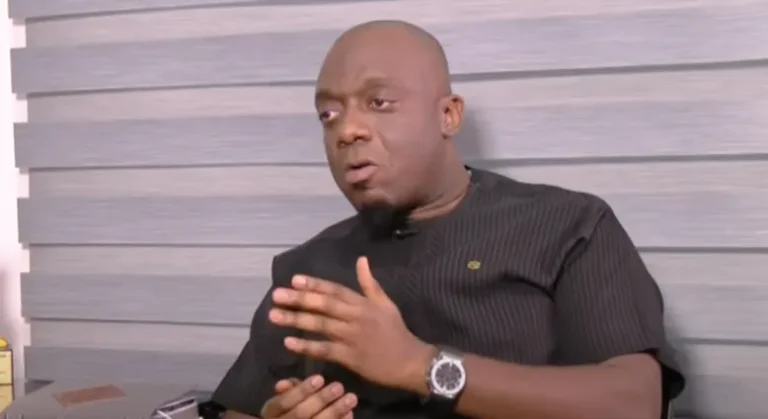 Man in a black shirt sits against a striped wall, gesturing with his hands as he speaks in an interview setting