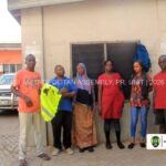 Seven people stand in a line outside a beige building, posing for a photo with a vehicle in the background; watermark reads Kumasi Metropolitan Assembly.