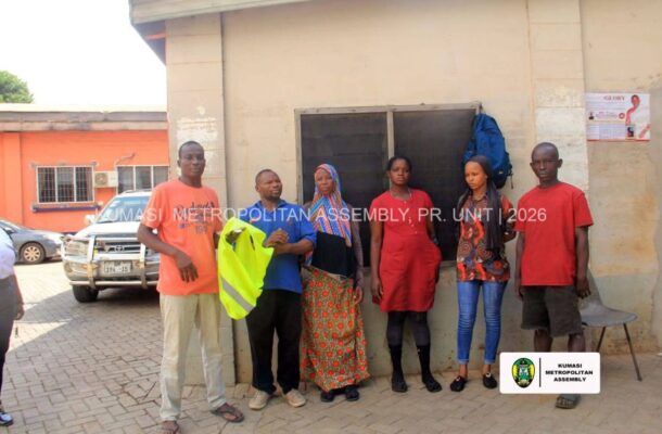 Seven people stand in a line outside a beige building, posing for a photo with a vehicle in the background; watermark reads Kumasi Metropolitan Assembly.