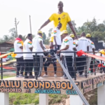 Group of people in white shirts gather on a decorated platform around a tall statue wearing a yellow shirt, at Kalili Roundabout with colorful ribbons and a crowd watching.
