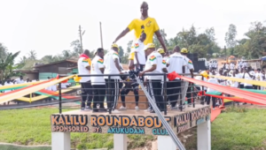 Group of people in white shirts gather on a decorated platform around a tall statue wearing a yellow shirt, at Kalili Roundabout with colorful ribbons and a crowd watching.
