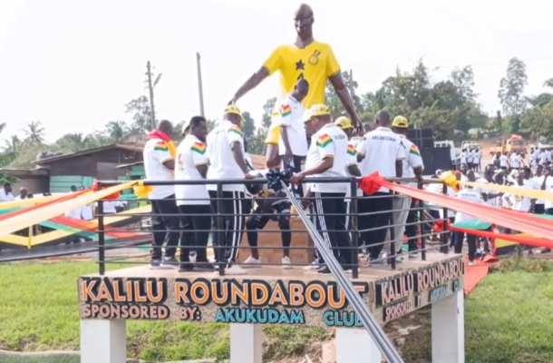 Group of people in white shirts gather on a decorated platform around a tall statue wearing a yellow shirt, at Kalili Roundabout with colorful ribbons and a crowd watching.