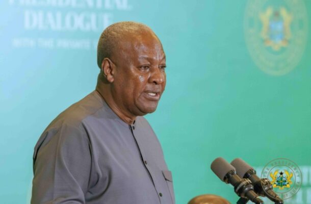Mahama-1-2-636x424-1 | Ghanaguardian.com Man in a gray shirt speaking at a press conference with multiple microphones at a podium, teal backdrop with government insignia behind him.