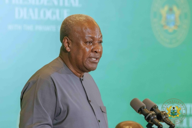 Man in a gray shirt speaking at a press conference with multiple microphones at a podium, teal backdrop with government insignia behind him.