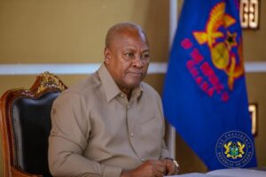 A man sits at a desk in a formal office, with a Ghana flag and the presidential seal visible in the background.