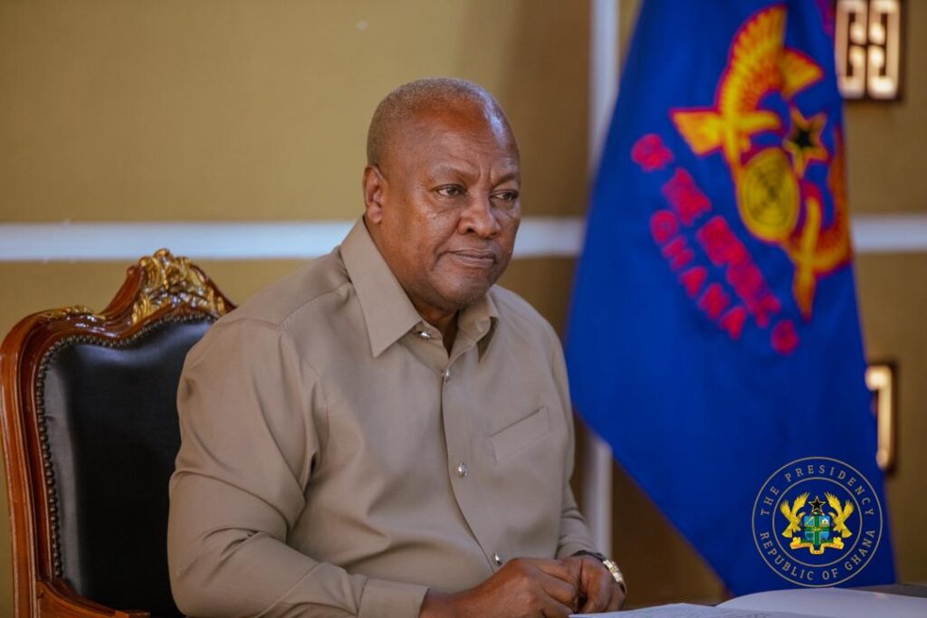 A man sits at a desk in a formal office, with a Ghana flag and the presidential seal visible in the background.