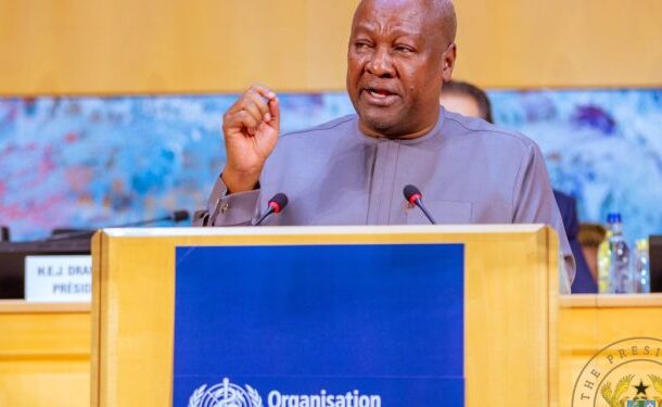 African man in a gray suit speaks at a podium with a blue UN banner, microphones, and officials in the background.