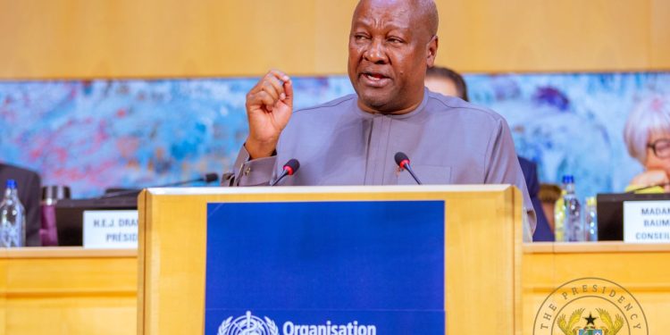 African man in a gray suit speaks at a podium with a blue UN banner, microphones, and officials in the background.
